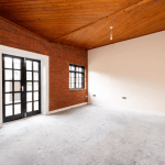 A spacious, unfurnished room with exposed red brick walls on the left and beige walls on the right. There's a pair of black-framed French doors and a window on the left wall. The ceiling is wooden, and the room is well-lit by natural light. The floor is carpeted—an ideal find at Palace Auctions.