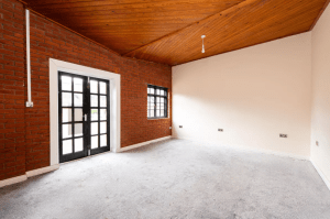A spacious, unfurnished room with exposed red brick walls on the left and beige walls on the right. There's a pair of black-framed French doors and a window on the left wall. The ceiling is wooden, and the room is well-lit by natural light. The floor is carpeted—an ideal find at Palace Auctions.