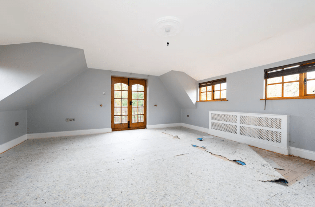 A spacious, unfurnished room with light blue walls, a light fixture on the ceiling, and a partially removed carpet exposing subflooring. The room features sloped ceilings, a pair of small windows to the right, and wooden double doors leading to a balcony straight ahead—a true gem in any Palace Auctions.