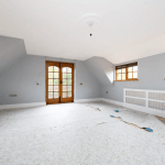 A spacious, unfurnished room with light blue walls, a light fixture on the ceiling, and a partially removed carpet exposing subflooring. The room features sloped ceilings, a pair of small windows to the right, and wooden double doors leading to a balcony straight ahead—a true gem in any Palace Auctions.