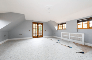 A spacious, unfurnished room with light blue walls, a light fixture on the ceiling, and a partially removed carpet exposing subflooring. The room features sloped ceilings, a pair of small windows to the right, and wooden double doors leading to a balcony straight ahead—a true gem in any Palace Auctions.