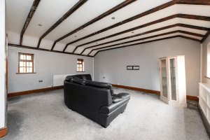 A sparsely furnished, spacious living room with exposed wooden ceiling beams, two small windows, a black leather couch, and a glass-fronted cabinet. The walls are light gray, and the floor is carpeted in a light color. This well-lit and airy space could grace any of the Cottages successfully sold at Palace Auctions.