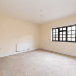 A vacant room with cream-colored walls and a beige carpet. The room has a large window with a black metal frame, allowing natural light to enter. There is a white radiator and some electrical outlets visible on the walls. Clean and spacious, the design could make this space highly sought-after at Palace Auctions.