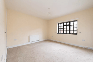 A vacant room with cream-colored walls and a beige carpet. The room has a large window with a black metal frame, allowing natural light to enter. There is a white radiator and some electrical outlets visible on the walls. Clean and spacious, the design could make this space highly sought-after at Palace Auctions.