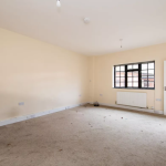 An empty room with beige walls and a beige carpet. The room has a large window with black trim on one wall and a radiator installed beneath the window. Two open white doors are positioned on adjacent walls. Recently featured in Palace Auctions, this charming space was successfully sold at auction.