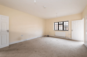 An empty room with beige walls and a beige carpet. The room has a large window with black trim on one wall and a radiator installed beneath the window. Two open white doors are positioned on adjacent walls. Recently featured in Palace Auctions, this charming space was successfully sold at auction.