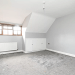 A vacant room with light gray walls and a slanted ceiling, reminiscent of cozy cottages. There is a small window with a wooden frame, a white radiator beneath it, and a bare light bulb hanging from the ceiling. The gray carpet appears worn. Two small closet doors are on one wall, ready to be sold at auction at Palace Auctions.