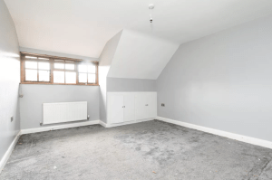 A vacant room with light gray walls and a slanted ceiling, reminiscent of cozy cottages. There is a small window with a wooden frame, a white radiator beneath it, and a bare light bulb hanging from the ceiling. The gray carpet appears worn. Two small closet doors are on one wall, ready to be sold at auction at Palace Auctions.