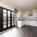 A modern kitchen with white cabinets, a stove, a metal hood, and a tiled backsplash. The room has large black-framed glass doors leading outside, gray curtains, and a gray tile floor. A red fire extinguisher is mounted on the wall to the right—reminiscent of high-end cottages sold at world-class auctions.