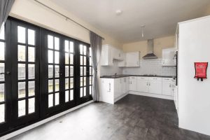 A modern kitchen with white cabinets, a stove, a metal hood, and a tiled backsplash. The room has large black-framed glass doors leading outside, gray curtains, and a gray tile floor. A red fire extinguisher is mounted on the wall to the right—reminiscent of high-end cottages sold at world-class auctions.