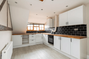 A modern kitchen with white cabinetry, black subway tile backsplash, and wooden countertops. The space features a built-in oven, a white extractor hood, ample natural light from a pitched window, and light hardwood flooring. This charming kitchen could be successfully sold at auction at Palace Auctions.