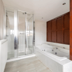 A modern bathroom in one of the POOLS COTTAGES, featuring a spacious glass-enclosed shower on the left and a large white bathtub with jets on the right. The room has light-colored walls and flooring, with wooden paneling on one side and a towel radiator near the shower.