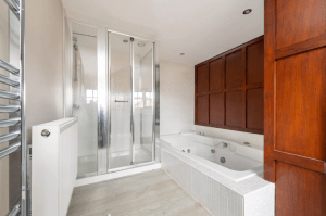 A modern bathroom in one of the POOLS COTTAGES, featuring a spacious glass-enclosed shower on the left and a large white bathtub with jets on the right. The room has light-colored walls and flooring, with wooden paneling on one side and a towel radiator near the shower.