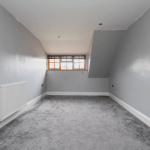 A small, empty room with slanted ceilings and gray-painted walls. The floor is covered with gray carpet. A single radiator is mounted on the left wall, and a large window with wooden blinds is centered on the back wall, allowing natural light to enter. Located in Kenilworth’s CV8 2JW and sold by auction with Palace Auctions.