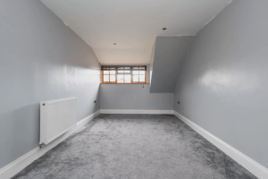 A small, empty room with slanted ceilings and gray-painted walls. The floor is covered with gray carpet. A single radiator is mounted on the left wall, and a large window with wooden blinds is centered on the back wall, allowing natural light to enter. Located in Kenilworth’s CV8 2JW and sold by auction with Palace Auctions.