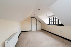 An empty attic room with sloped ceilings, beige walls, and a carpeted floor. There are recessed ceiling lights, a door in the back wall, a radiator on the left wall, and a window on the right wall. Located at Kenilworth, CV8 2JW and sold by auction with Palace Auctions.