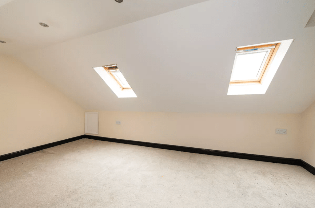A small attic room with sloped ceilings features two skylight windows allowing natural light to flood in. The room, located in the charming Pools Cottages, Kenilworth, has beige walls, a light-colored carpet, and recessed lighting on the ceiling. This delightful space was CV8 2JW sold by auction with Palace Auctions.
