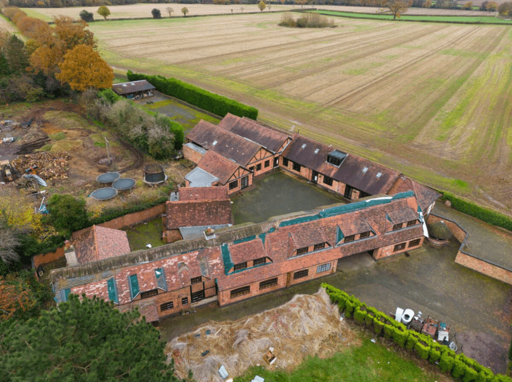 Aerial view of a large, U-shaped brick building complex with multiple pitched roofs, surrounded by fields and trees in Kenilworth, CV8 2JW. The compound includes several small structures and enclosed areas, with green hedges and scattered equipment visible around the perimeter. Sold by Auction with Palace Auctions.