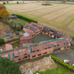 Aerial view of a large, U-shaped brick building complex with multiple pitched roofs, surrounded by fields and trees in Kenilworth, CV8 2JW. The compound includes several small structures and enclosed areas, with green hedges and scattered equipment visible around the perimeter. Sold by Auction with Palace Auctions.