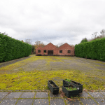 A spacious outdoor area with a moss-covered ground leads to two small red-brick buildings at the center, known as POOLS COTTAGES in Kenilworth. The perimeter is bordered by tall, dense green hedges. In the foreground, there are two black planters: one upright with few plants and one fallen over. CV8 2JW Sold by Auction with Palace Auctions.