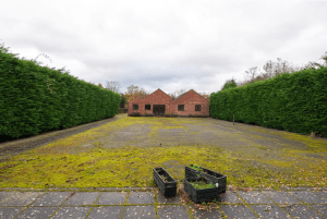 A spacious outdoor area with a moss-covered ground leads to two small red-brick buildings at the center, known as POOLS COTTAGES in Kenilworth. The perimeter is bordered by tall, dense green hedges. In the foreground, there are two black planters: one upright with few plants and one fallen over. CV8 2JW Sold by Auction with Palace Auctions.