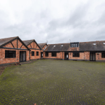 A large courtyard surrounded by one-story brick cottages with multiple windows and dark framed doors. The pavement of the courtyard is a mix of patches of green moss and a herringbone brick pattern. The sky is overcast, adding to the subdued atmosphere in Kenilworth, CV8 2JW Sold by Auction with Palace Auctions.