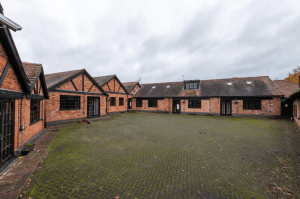 A large courtyard surrounded by one-story brick cottages with multiple windows and dark framed doors. The pavement of the courtyard is a mix of patches of green moss and a herringbone brick pattern. The sky is overcast, adding to the subdued atmosphere in Kenilworth, CV8 2JW Sold by Auction with Palace Auctions.