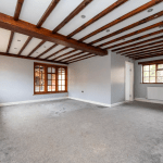 A spacious room with light gray walls and carpet, reminiscent of the charm found in cottages successfully sold at auction. The room features wooden ceiling beams and windows on three walls, allowing natural light to brighten the space. There are recessed lights in the ceiling, and a radiator is visible under one of the windows.