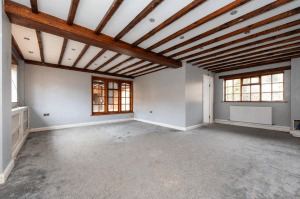 A spacious room with light gray walls and carpet, reminiscent of the charm found in cottages successfully sold at auction. The room features wooden ceiling beams and windows on three walls, allowing natural light to brighten the space. There are recessed lights in the ceiling, and a radiator is visible under one of the windows.