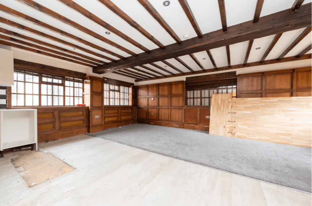 A large, empty room with exposed wooden ceiling beams, wood-paneled walls, and large windows. The space has a mix of carpeted and bare floors, with some construction materials like loose boards leaning against the wall. Recently featured in Cottages successfully sold at auction by Palace Auctions.