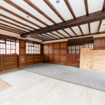 A large, empty room with exposed wooden ceiling beams, wood-paneled walls, and large windows. The space has a mix of carpeted and bare floors, with some construction materials like loose boards leaning against the wall. Recently featured in Cottages successfully sold at auction by Palace Auctions.
