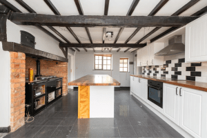 A spacious kitchen featuring black ceiling beams, white cabinets, and black countertops. The room has a black and white tile backsplash and a center island with a wood countertop. An exposed brick wall contains a black stove, and the kitchen has a modern, minimalistic style reminiscent of auctioned cottages.
