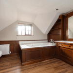 An attic bathroom with a wooden bathtub surround and matching cabinetry under a large mirror exudes rustic charm. The room features sloped ceilings, a small window with blinds, and a white toilet next to a radiator. With its wooden flooring, this cottage-style oasis feels like it could be successfully sold at auction.