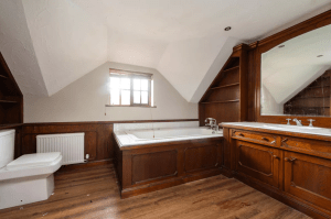 An attic bathroom with a wooden bathtub surround and matching cabinetry under a large mirror exudes rustic charm. The room features sloped ceilings, a small window with blinds, and a white toilet next to a radiator. With its wooden flooring, this cottage-style oasis feels like it could be successfully sold at auction.