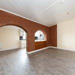 An empty room with brick archways leading to a kitchen area. The room has beige walls, a white ceiling, and grey wood flooring. The kitchen features partial black and white tiled walls and wooden cabinets. A radiator is visible in the adjacent area. Part of Pools Cottages, it was successfully sold at Palace Auctions.