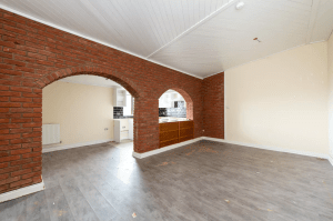 An empty room with brick archways leading to a kitchen area. The room has beige walls, a white ceiling, and grey wood flooring. The kitchen features partial black and white tiled walls and wooden cabinets. A radiator is visible in the adjacent area. Part of Pools Cottages, it was successfully sold at Palace Auctions.