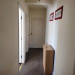 A narrow hallway with white walls and a carpeted floor. There is a door at the end of the hallway and an open door on the left side. Two red-framed pictures hang on the right wall, and a small wooden cabinet with a few items sits beneath them in what was an apartment in Samuels Tower, successfully sold at Palace Auctions.
