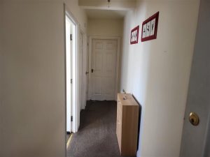 A narrow hallway with white walls and a carpeted floor. There is a door at the end of the hallway and an open door on the left side. Two red-framed pictures hang on the right wall, and a small wooden cabinet with a few items sits beneath them in what was an apartment in Samuels Tower, successfully sold at Palace Auctions.