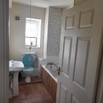 A small bathroom in the recently sold apartment in Samuels Tower features a white sink and toilet with a blue toilet seat. A rectangular bathtub is adjacent to the toilet, with a white and gray tiled wall above it. Natural light flows through a small window above the toilet, and the door is open.