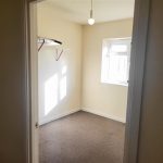 A small, empty room with beige walls and a bare light bulb hanging from the ceiling. There is a window on the right side letting in sunlight, illuminating part of the room. A shelf with a hanging bar is installed on the left wall. The carpeted floor reflects its recent success at auction in Samuels Tower, sold at Palace Auctions.