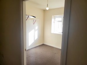A small, empty room with beige walls and a bare light bulb hanging from the ceiling. There is a window on the right side letting in sunlight, illuminating part of the room. A shelf with a hanging bar is installed on the left wall. The carpeted floor reflects its recent success at auction in Samuels Tower, sold at Palace Auctions.