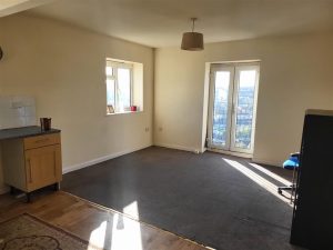 A sparsely furnished apartment in Samuels Tower boasts beige walls and a dark carpet. A small wooden cabinet on the left, a dark grey shelf on the right, and sunlight streaming through glass doors reveal a cityscape view. This stunning space was successfully sold at auction at Palace Auctions.