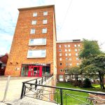 A multi-story red-brick building with numerous windows and red-framed entrance doors stands proudly, surrounded by a neatly maintained lawn with trees and shrubs. Adjacent to the main building are smaller brick structures. The sky is partly cloudy—a picturesque view from New Alexander Court Nottingham, where Palace Auctions has two investment apartments for sale by auction.
