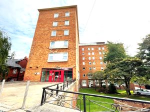 A multi-story red-brick building with numerous windows and red-framed entrance doors stands proudly, surrounded by a neatly maintained lawn with trees and shrubs. Adjacent to the main building are smaller brick structures. The sky is partly cloudy—a picturesque view from New Alexander Court Nottingham, where Palace Auctions has two investment apartments for sale by auction.