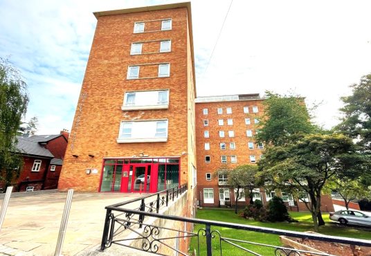 A multi-story red-brick building with numerous windows and red-framed entrance doors stands proudly, surrounded by a neatly maintained lawn with trees and shrubs. Adjacent to the main building are smaller brick structures. The sky is partly cloudy—a picturesque view from New Alexander Court Nottingham, where Palace Auctions has two investment apartments for sale by auction.