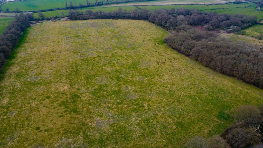 Aerial view of a large grassy field surrounded by wooded areas and farmland, reflecting parcels of premodernity agricultural land. The grass shows varying stages of growth with patches of green and brown. Dense trees border the field, where faint trails are visible. Available for sale by auction with Palace Auctions.