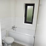 A small bathroom with white tiled walls and a frosted window offers views of Ruth House. It features a white bathtub with a silver faucet, a white toilet, and beige tiled flooring. The door is partially open. Available for sale by auction with Palace Auctions.