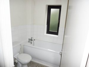 A small bathroom with white tiled walls and a frosted window offers views of Ruth House. It features a white bathtub with a silver faucet, a white toilet, and beige tiled flooring. The door is partially open. Available for sale by auction with Palace Auctions.