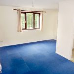 A bright, empty room features a window with beige curtains, white walls, and a clean blue carpet. The space is well-lit by natural light from the window, offering views of Ruth House. An adjacent kitchen is visible through an open doorway, with light-colored tile flooring. Available for sale by auction with Palace Auctions.