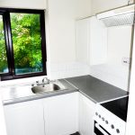 A compact kitchen with white cabinetry and countertops. A sink is beneath a window with a view of green foliage and glimpses of the historic Ruth House. A stove with an extractor fan is to the right. The backsplash is tiled in white, and the floor has gray tiles. Available for sale by auction with Palace Auctions.